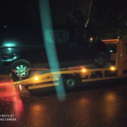 GITCARS Izabela Chruściel - Ciemny bus zabezpieczony pasami na żółtej lawecie, sfotografowany nocą na mokrej nawierzchni.