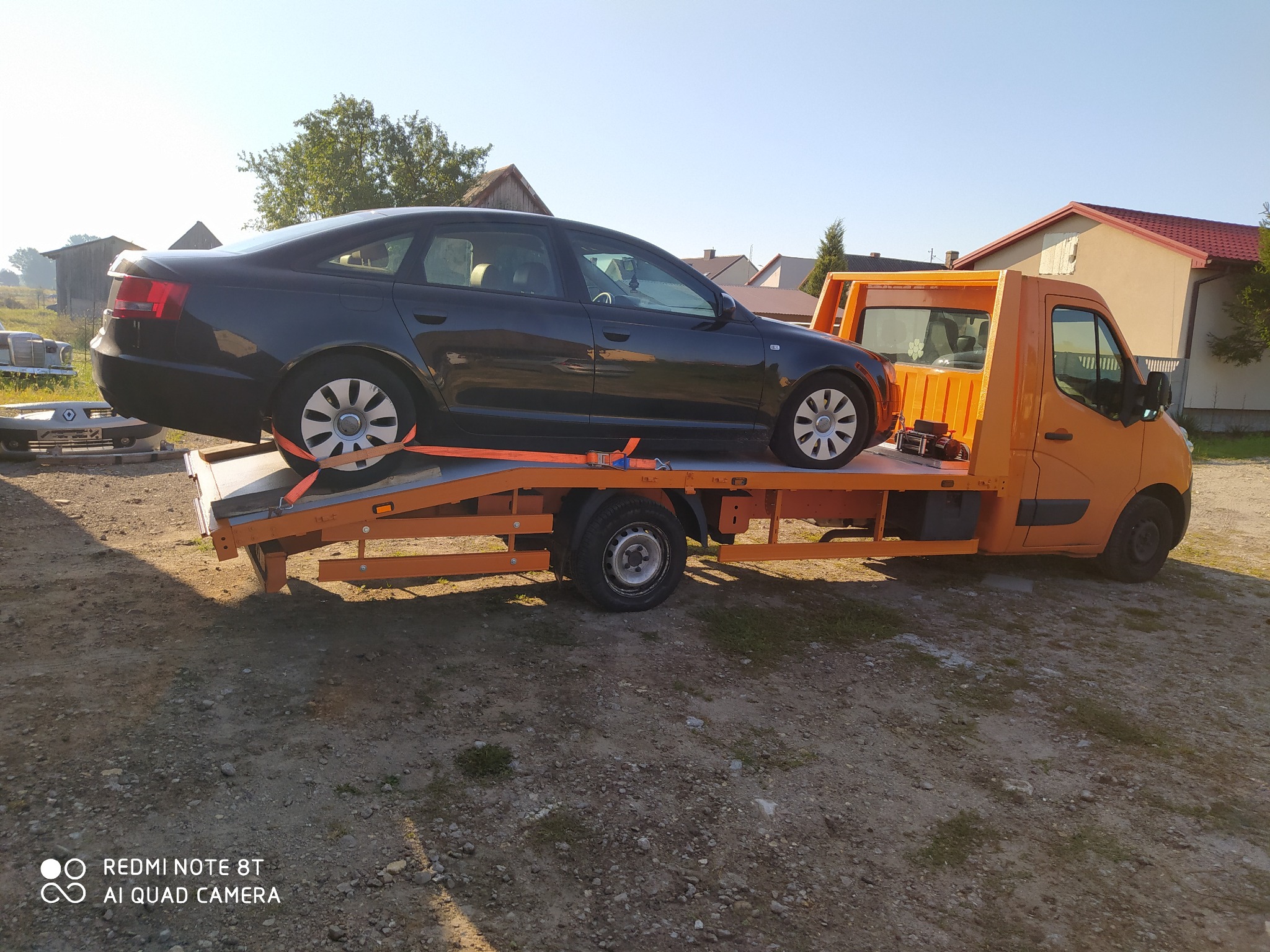 Czarny samochód osobowy zabezpieczony pomarańczowymi pasami na pomarańczowej lawecie, w tle zabudowania gospodarcze i drzewa.