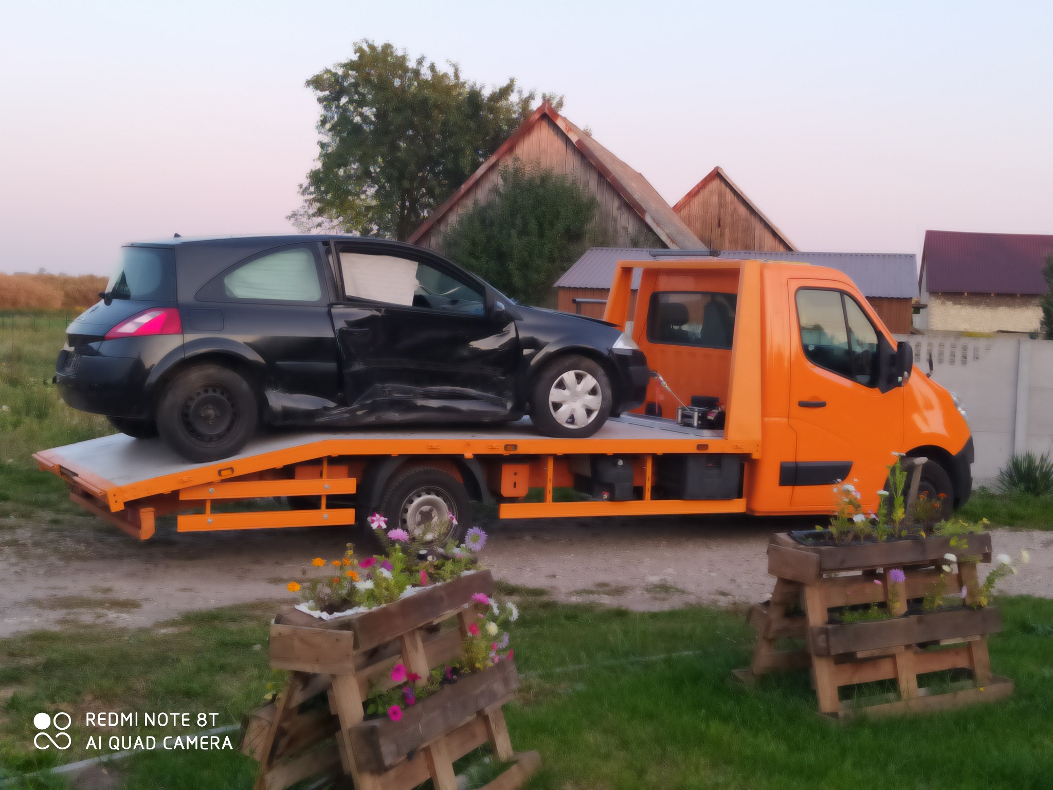 Pomarańczowa laweta z uszkodzonym czarnym samochodem na platformie, na tle wiejskich zabudowań i zachodzącego słońca, z drewnianą konstrukcją z kwiatami na pierwszym planie.