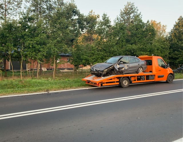 Pomarańczowa laweta z uszkodzonym szarym samochodem osobowym na platformie, widok z boku na tle drzew i drogi.