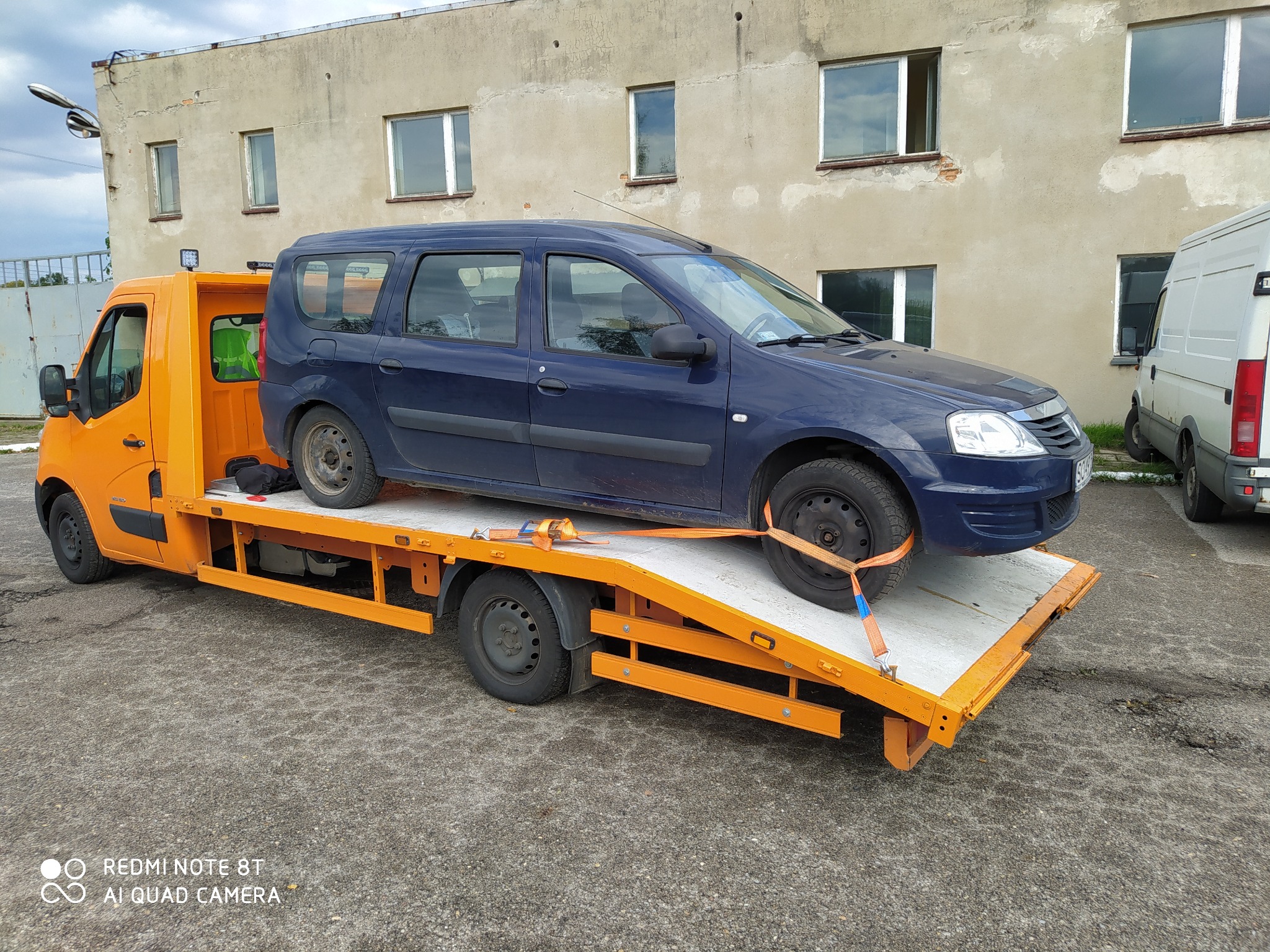 Pomarańczowa laweta z niebieskim kombi zabezpieczonym pasami transportowymi na platformie, na tle budynku z oknami.