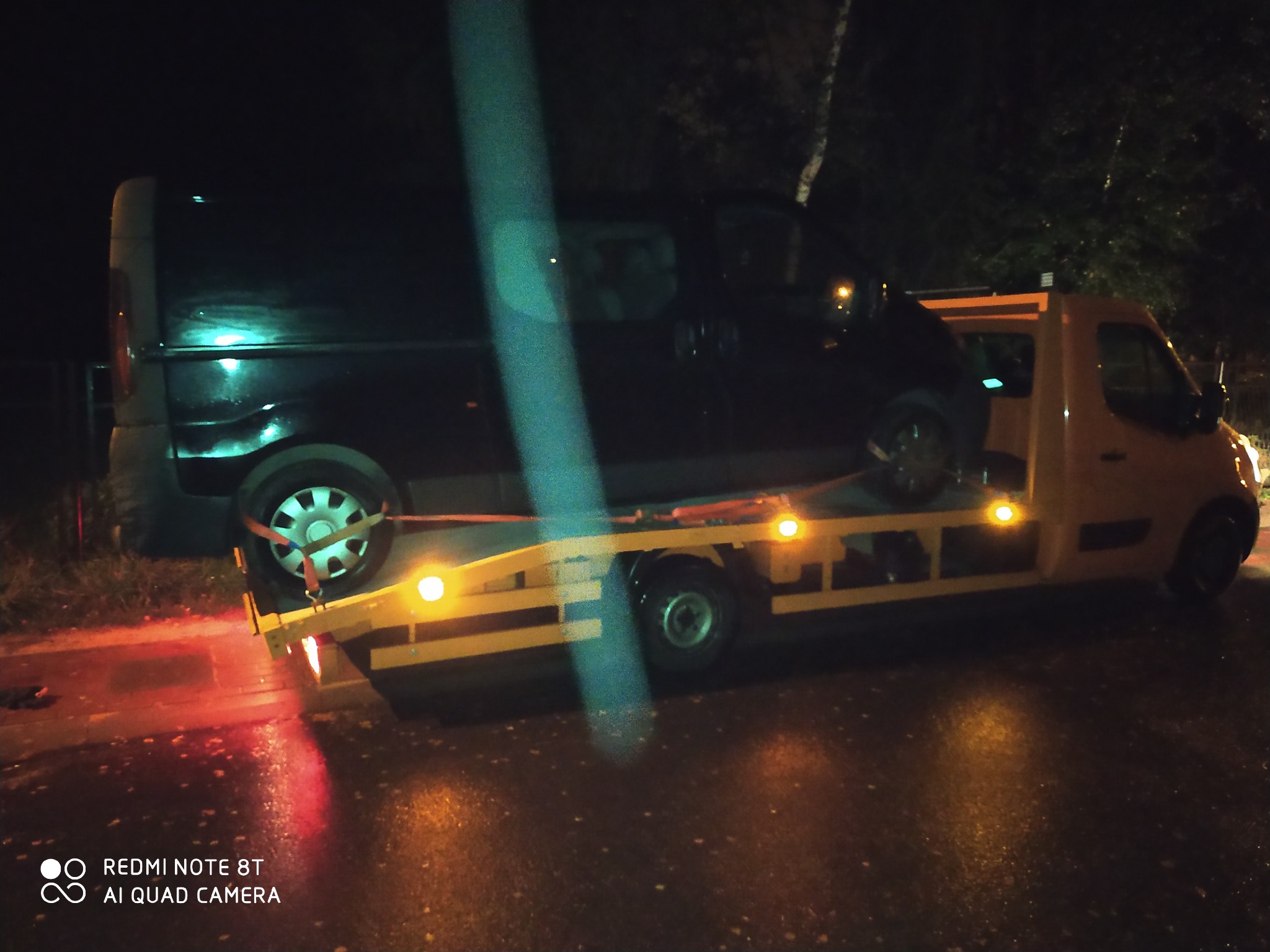 Ciemny bus zabezpieczony pasami na żółtej lawecie, sfotografowany nocą na mokrej nawierzchni.