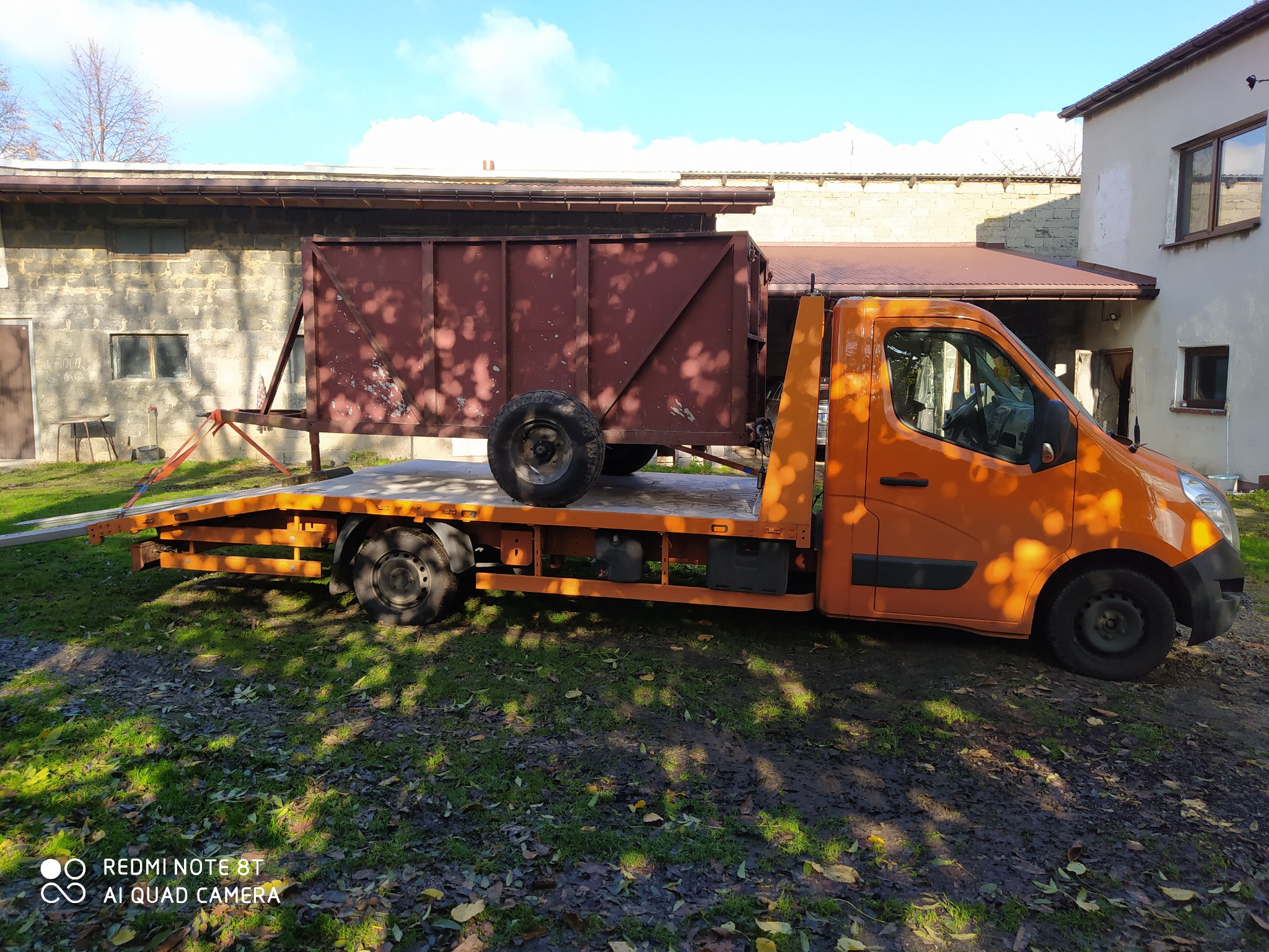 Pomarańczowa laweta z przyczepą skrzyniową na platformie, zaparkowana na trawiastym terenie przed budynkiem z cegły.