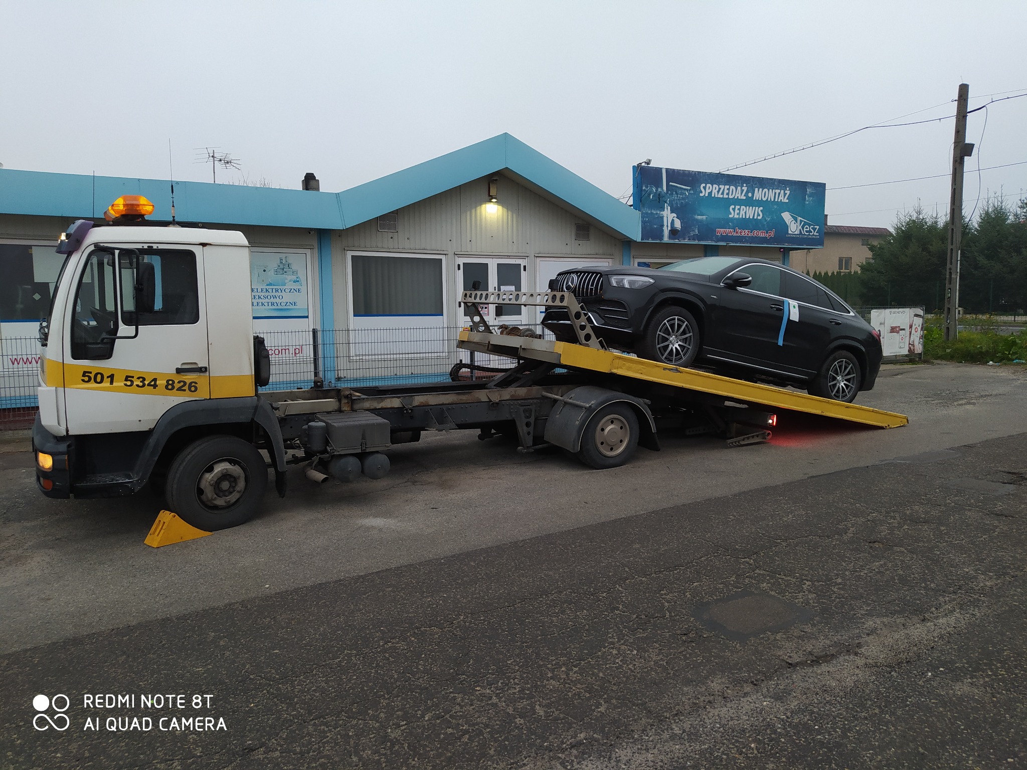 Czarny samochód osobowy marki Mercedes transportowany na żółtej lawecie w Przyrowie, widok z boku, w tle budynek z reklamą.