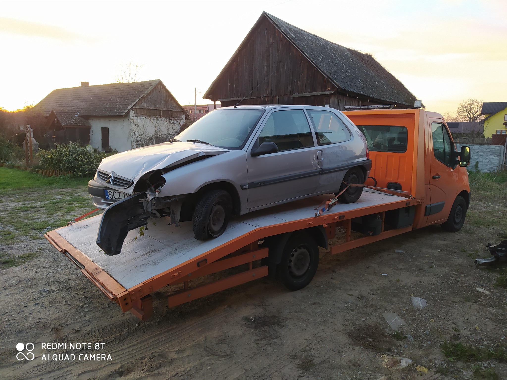 Uszkodzony srebrny samochód osobowy marki Citroen, z widocznymi uszkodzeniami przodu, załadowany na pomarańczową platformę holowniczą w wiejskim otoczeniu.