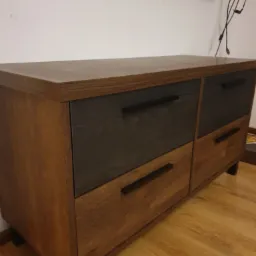 Komoda z drewnianym korpusem w odcieniach brązu i szarymi frontami szuflad, wyposażona w czarne, podłużne uchwyty, ustawiona na podłodze z paneli.