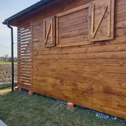 Zewnętrzny widok nowej, drewnianej konstrukcji z oknami z okiennicami, pomalowanej na brązowo, z elementami ażurowymi po lewej stronie, osadzonej na betonowych podstawach.