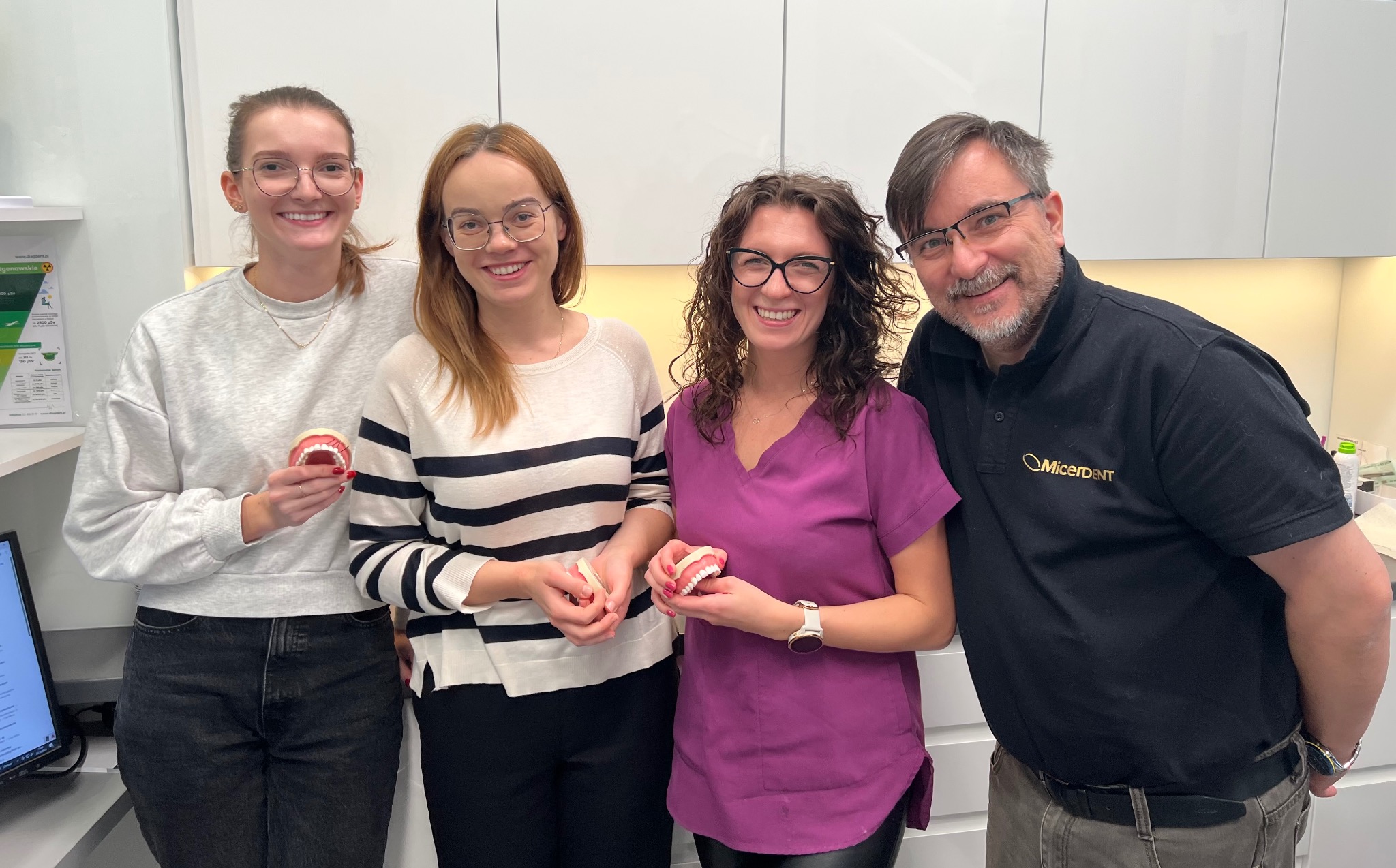 Zespół stomatologów prezentuje modele szczęk w gabinecie. Widoczny mężczyzna w koszulce z logo MicerDent. Jasne wnętrze, uśmiechnięte twarze.