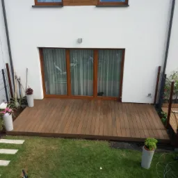 Widok z góry na taras z desek w kolorze ciepłego brązu, przylegający do białego domu z trzema oknami tarasowymi; w oknach widoczne zasłony, na tarasie cień kota, obok tarasu trawnik z betonowymi...