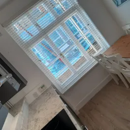 Ukośne ujęcie jasnej kuchni z drewnianą podłogą, szarą zabudową, marmurowym blatem i drewnianym stołem z białymi krzesłami, widok przez żaluzje na zewnątrz.