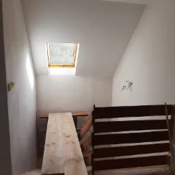 Pokój na poddaszu w trakcie wykańczania, widoczne zabezpieczone folią podłogi, surowe ściany, okno dachowe i przygotowane deski na rusztowaniu.