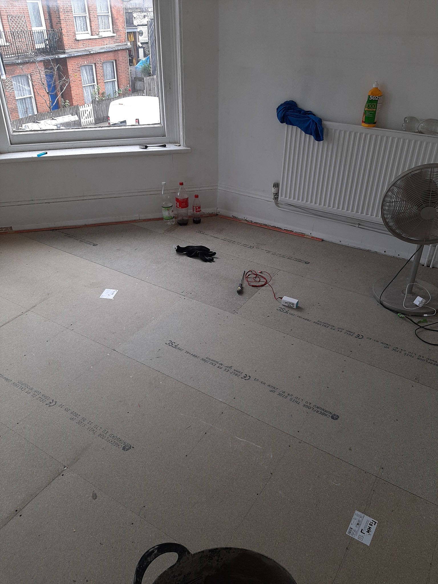 Przygotowana podłoga z płyt podkładowych pod panele lub parkiet, widoczne narzędzia pomiarowe, butelki z napojami i rękawice robocze, okno z widokiem na budynek mieszkalny.