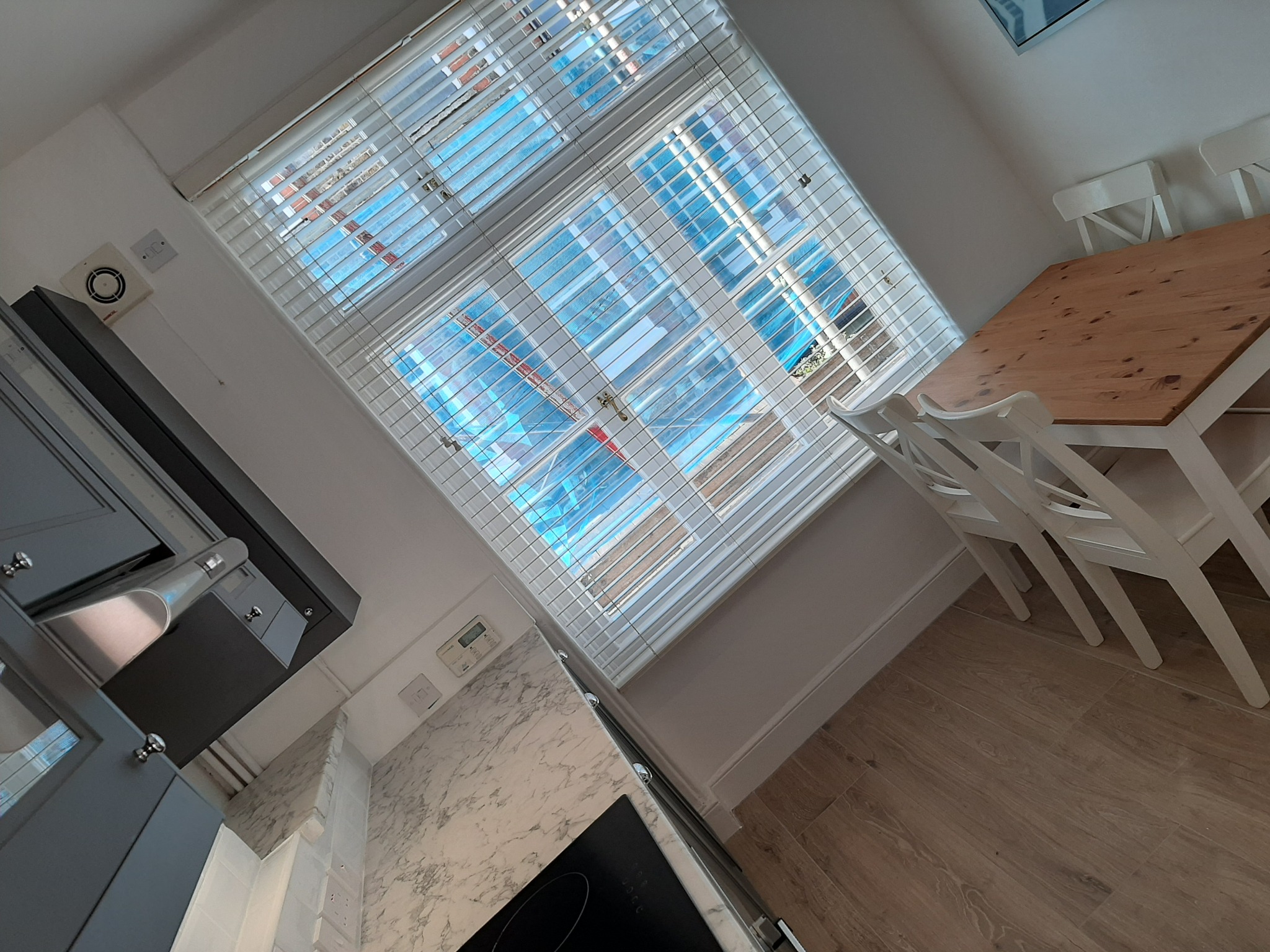 Ukośne ujęcie jasnej kuchni z drewnianą podłogą, szarą zabudową, marmurowym blatem i drewnianym stołem z białymi krzesłami, widok przez żaluzje na zewnątrz.