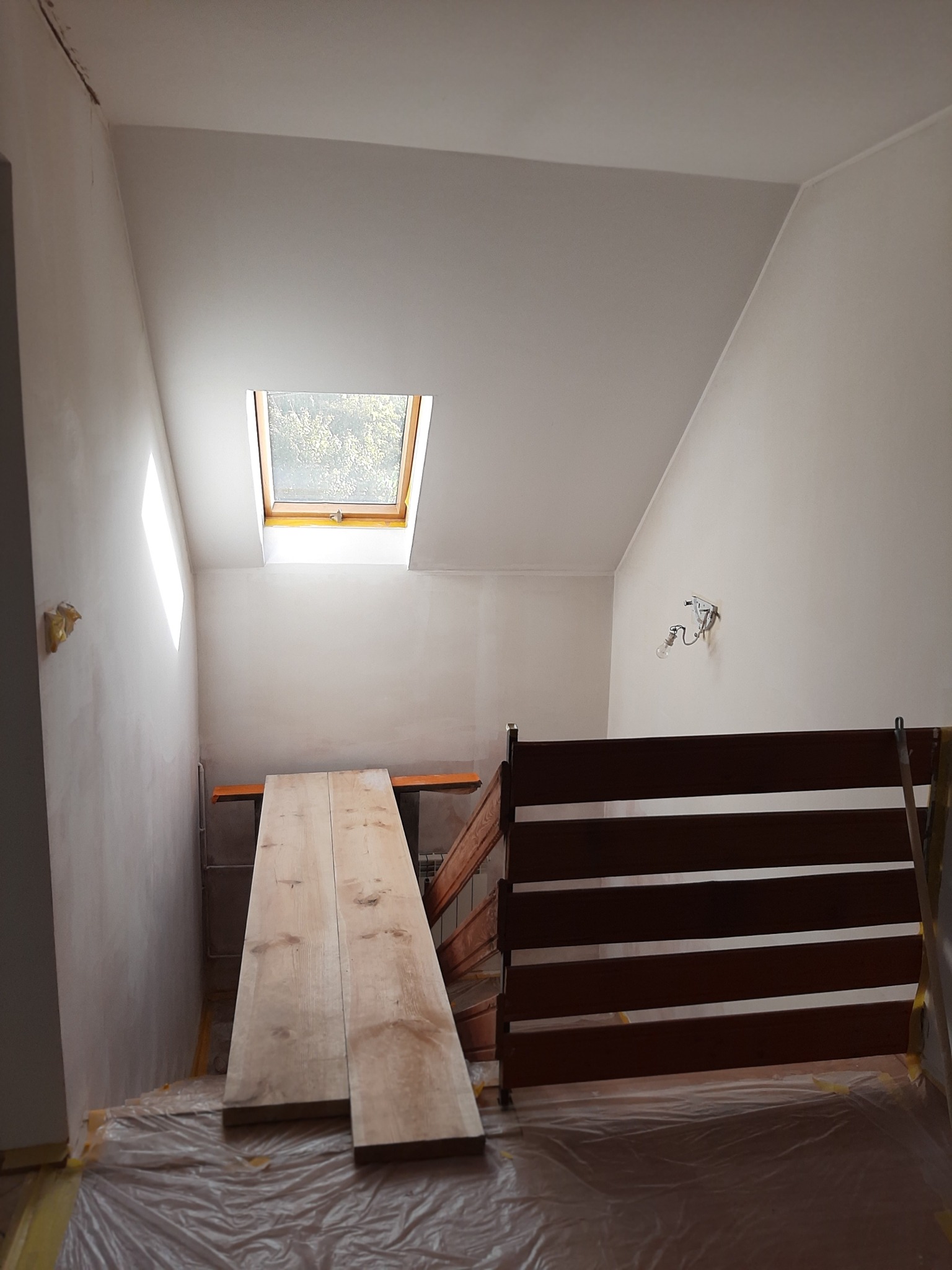 Pokój na poddaszu w trakcie wykańczania, widoczne zabezpieczone folią podłogi, surowe ściany, okno dachowe i przygotowane deski na rusztowaniu.