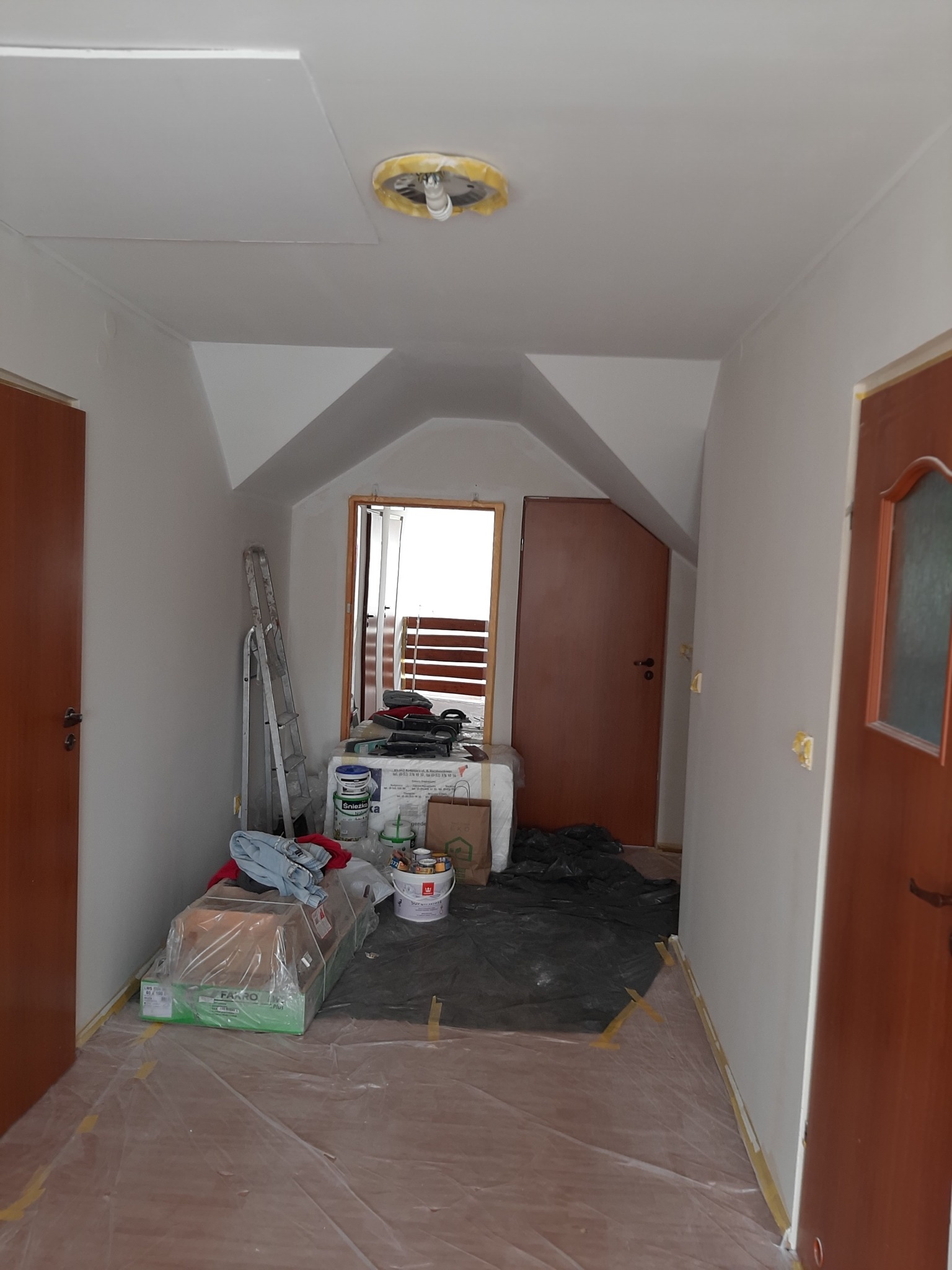 Przygotowania do malowania korytarza z dwoma parami drzwi, zabezpieczona podłoga folią, drabina oparta o ścianę, puszki z farbą i narzędzia na podłodze.