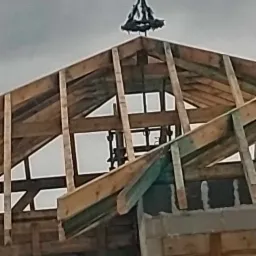 Konstrukcja drewniana dachu w trakcie budowy, widoczna więźba dachowa z elementami dekoracyjnymi na szczycie, pochmurne niebo w tle.