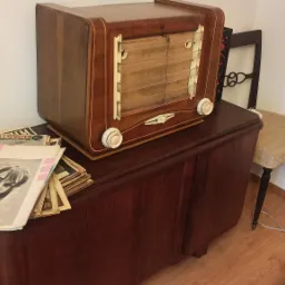 Antyczne radio stojące na odrestaurowanej komodzie, obok stos starych magazynów i krzesła