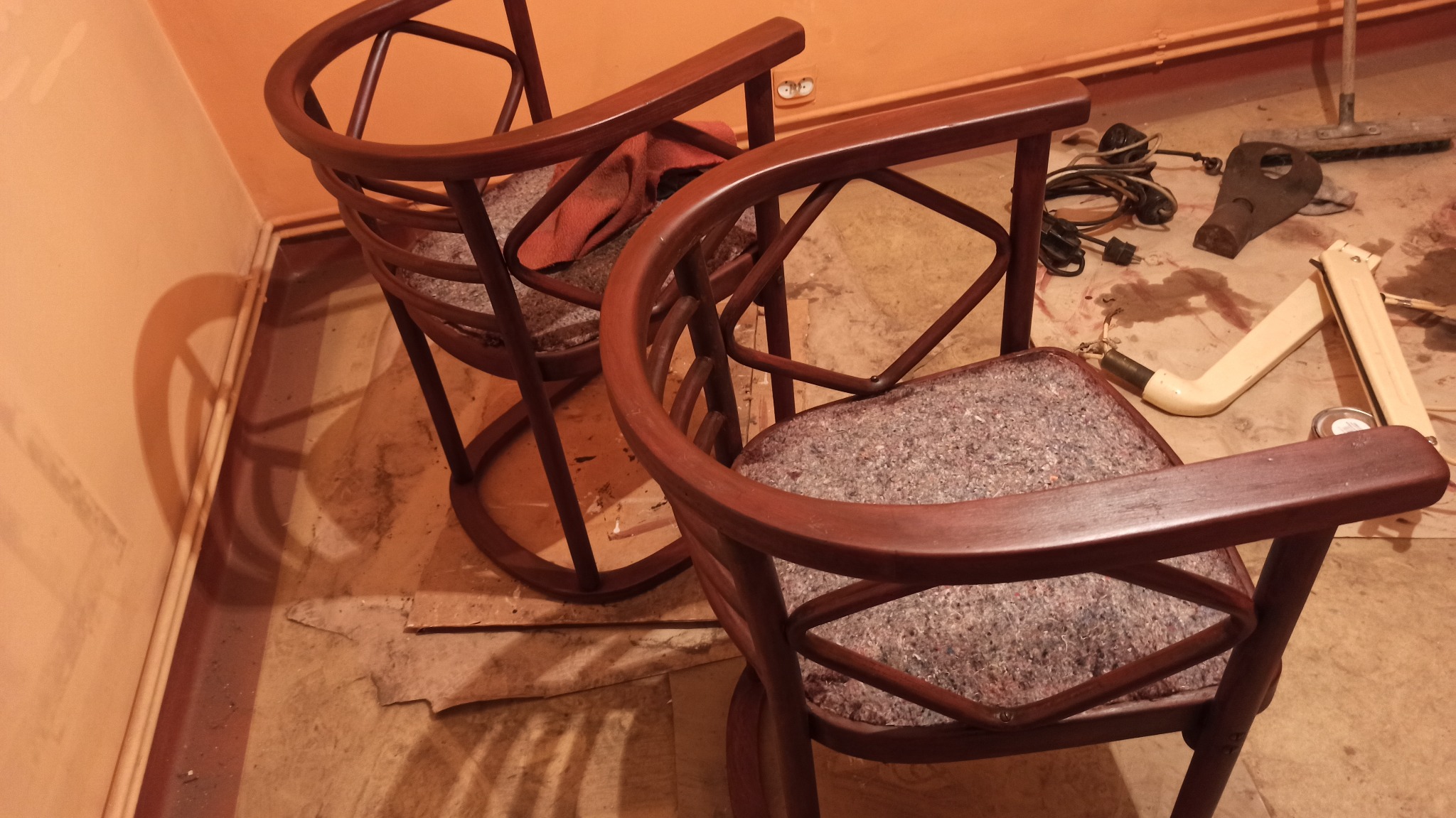 Dwa krzesła z ciemnego drewna z szarymi siedziskami na podłodze w trakcie renowacji, obok narzędzia i akcesoria.