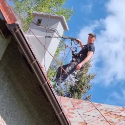 Pracownik w uprzęży alpinistycznej myje dach myjką ciśnieniową, widoczny komin, dachówka i niebieskie niebo.
