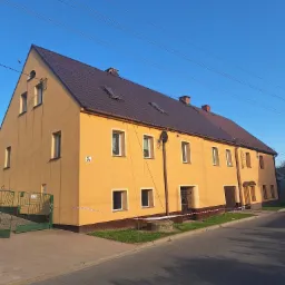 Odnowiony budynek wielorodzinny z brązowym dachem, pomalowany na żółto, z widoczną elewacją i rynnami, zabezpieczony taśmą ostrzegawczą.