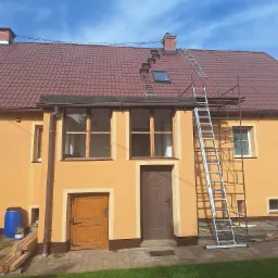 Dwupiętrowy dom z pomarańczową elewacją i czerwoną dachówką, z rozstawioną drabiną na dachu i rusztowaniem przy ścianie, sugerujące prace remontowe lub konserwacyjne.