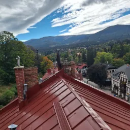 Czerwony dach z blachy na tle górskiego krajobrazu i zabudowy miejskiej, widoczne kominy i metalowy stopień dachowy.