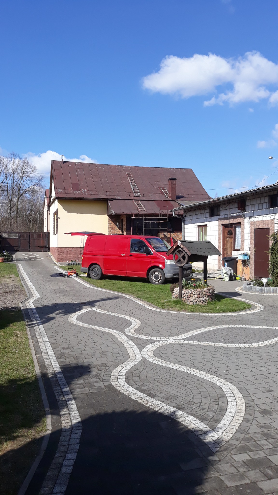 Czerwony samochód dostawczy zaparkowany przed domem z rusztowaniem przy dachu, studnia z kamienną obudową na pierwszym planie.