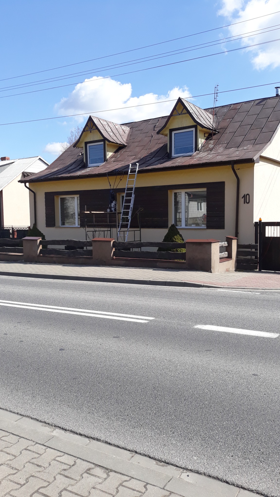 Dwukondygnacyjny dom z brązowym dachem i beżową elewacją, z drabina opartą o dach, widoczne ślady zabrudzeń na dachu, ujęcie z poziomu ulicy.