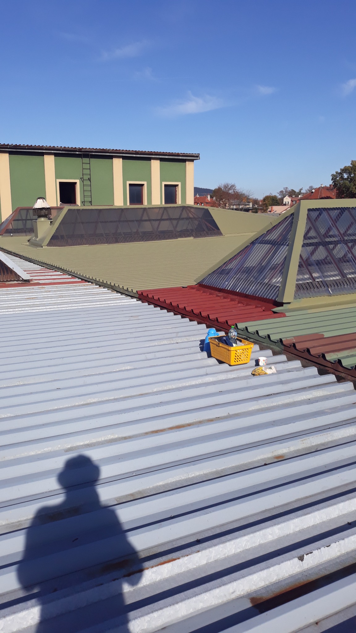 Widok z góry na blaszany dach z koszykiem narzędzi i cieniem osoby, na tle zielono-beżowego budynku z oknami i świetlikami dachowymi.