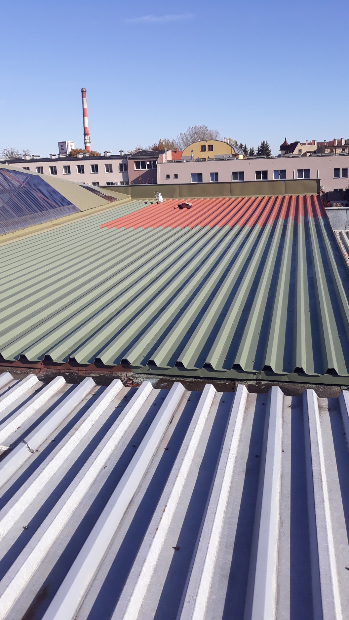 Częściowo pomalowany blaszany dach w kolorze czerwonym, z puszką farby i pędzlem, widok z góry na budynki i komin w tle.