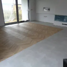 Fragment wnętrza w trakcie wykańczania: ułożona część podłogi z paneli w jodełkę kontrastuje z surową wylewką, okno zabezpieczone folią.