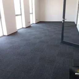 Wnętrze biurowe z ciemnoszarą wykładziną dywanową w kwadraty, widoczne okna i otwarte drzwi wejściowe, logo firmy w rogu.