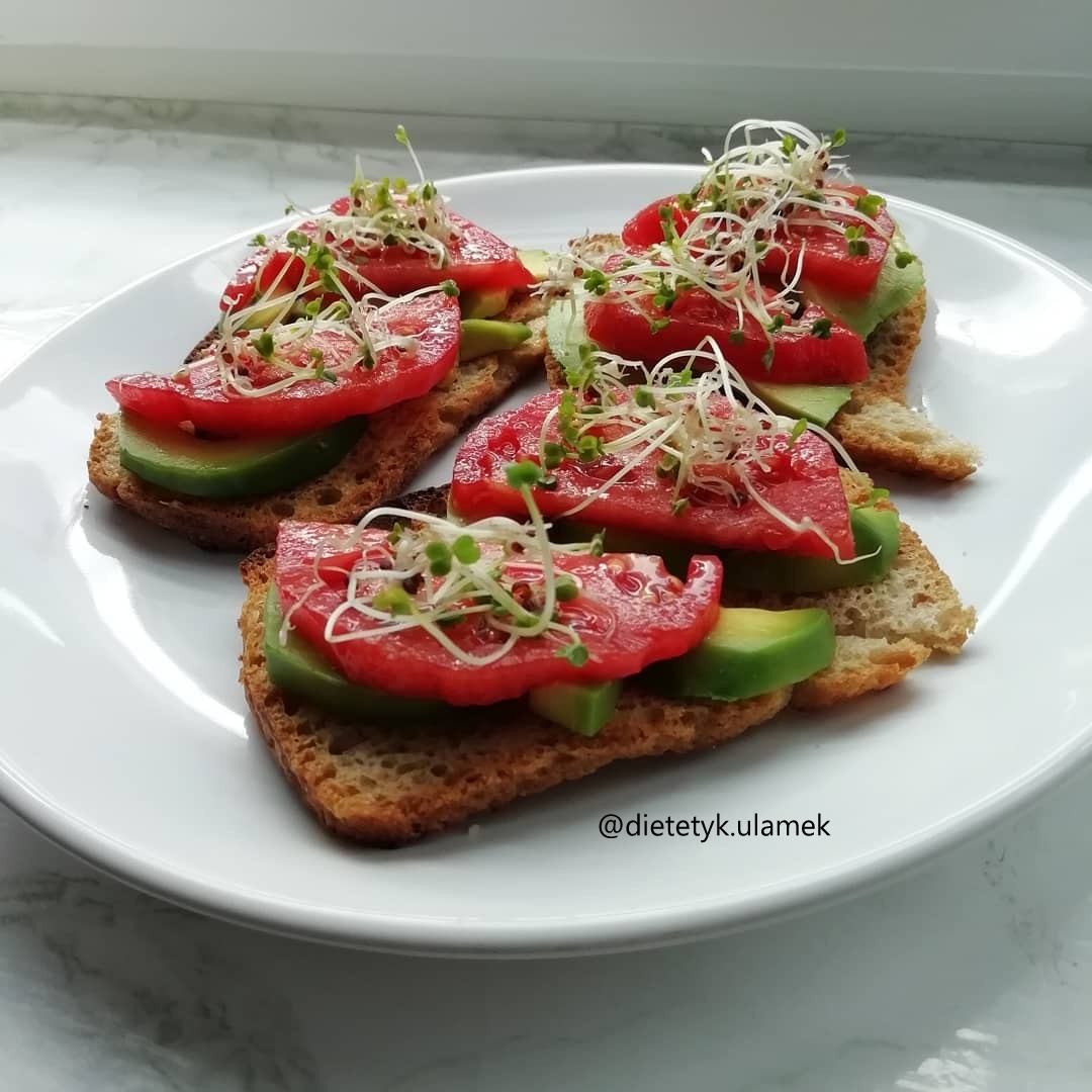 Cztery kanapki z chleba tostowego, awokado, pomidorami i kiełkami na białym talerzu na tle marmurowego blatu.