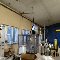 Wnętrze hali produkcyjnej z maszynami przemysłowymi, kartonami i instalacjami pod sufitem.