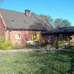 Ceglany dom jednorodzinny z brązowym dachem i żółtymi okiennicami w Bielsko-Białej, otoczony zielonym trawnikiem z opadłymi liśćmi i rabatami kwiatowymi.