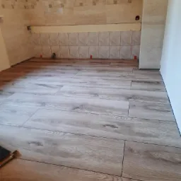 Jasne panele podłogowe imitujące drewno, ułożone w nowej łazience z beżowymi płytkami na ścianach, widoczne elementy montażowe i niewielki fragment paczki paneli.