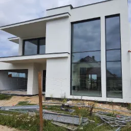 Nowoczesny, dwukondygnacyjny dom z białymi ścianami i dużymi oknami z czarnymi ramami, częściowo otoczony siatką i rusztowaniami, sugerujący prace wykończeniowe elewacji.