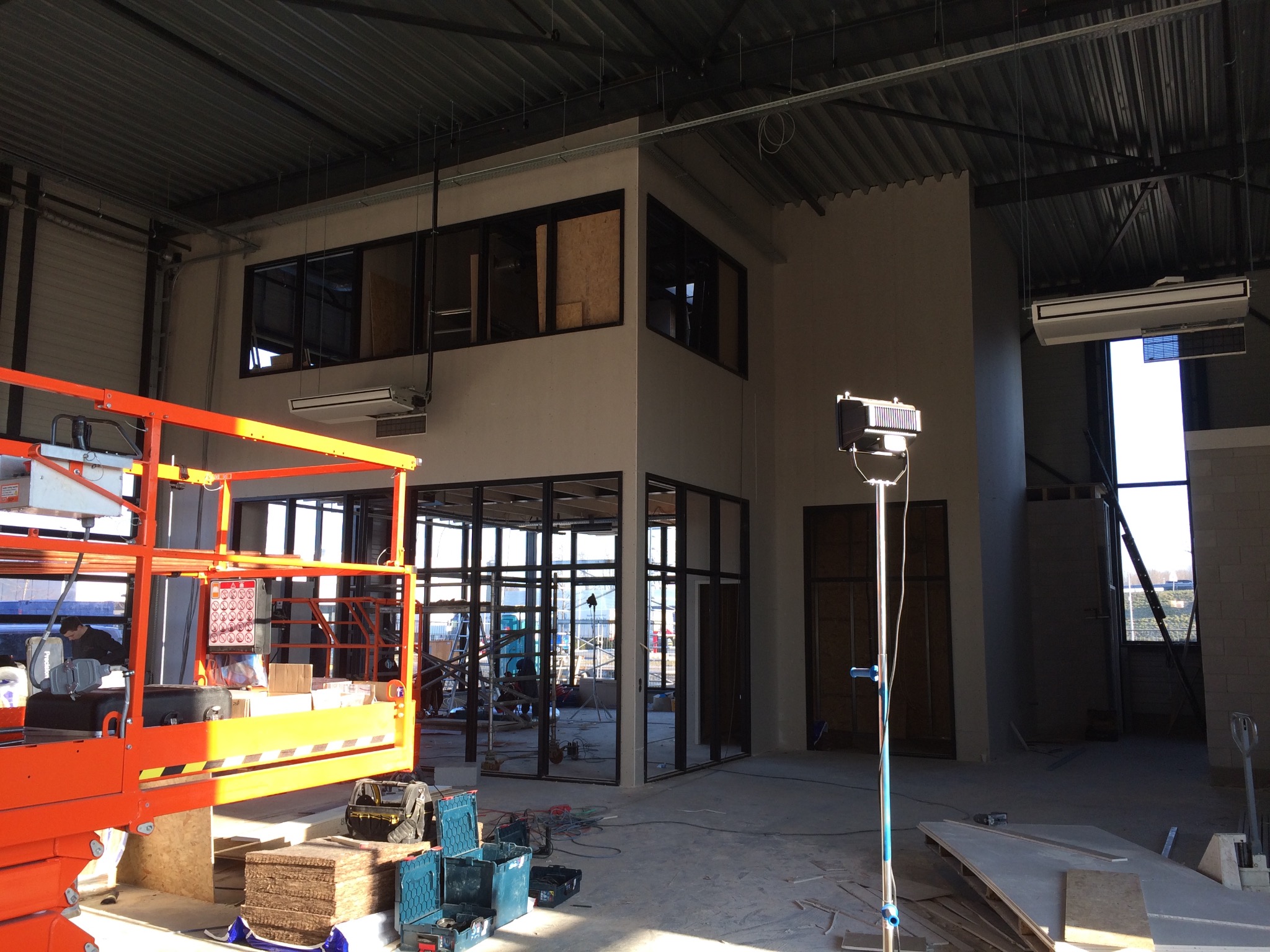 Wnętrze biura w stanie surowym podczas prac wykończeniowych: widoczna pomarańczowa platforma robocza, narzędzia, metalowa konstrukcja ścian działowych z czarnymi ramami okiennymi, oświetlenie...