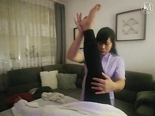 Terapeuta w jasnofioletowej koszuli rozciąga nogę klienta leżącego na stole do masażu w pokoju z szarym narożnikiem i zasłonami w tle.