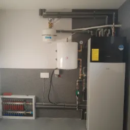 Nowoczesna instalacja grzewcza z pompą ciepła Daikin, zasobnikiem ciepłej wody i rozdzielaczem podłogówki w kotłowni wykończonej szarą mozaiką.