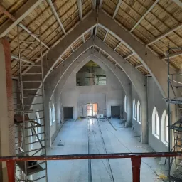 Wnętrze remontowanego budynku z wysokim sklepieniem łukowym, widoczne rusztowania i surowy stan wykończenia ścian oraz drewniany strop