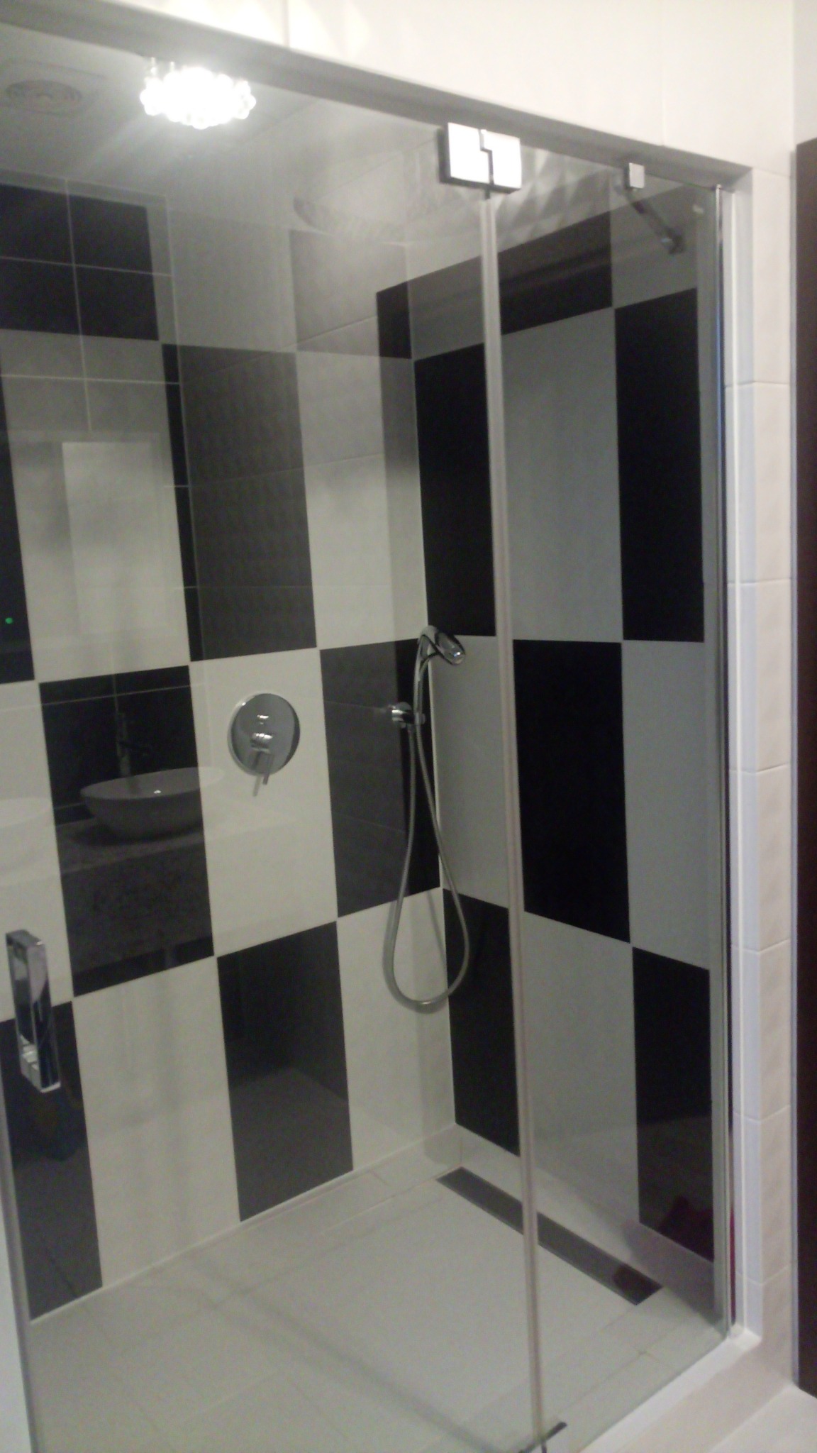 Kabina prysznicowa z czarno-białymi płytkami w szachownicę, chromowana armatura i odpływ liniowy.