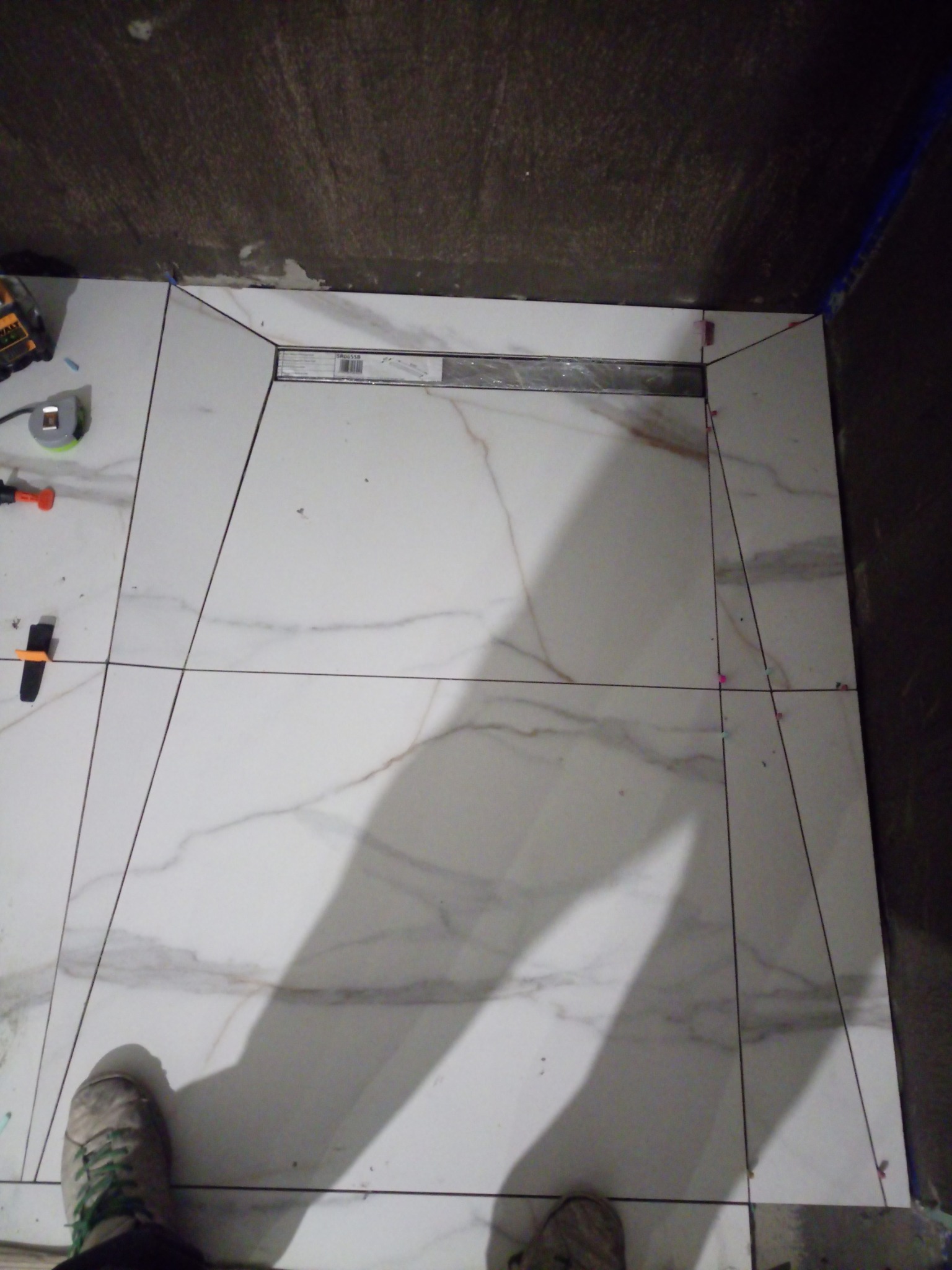 Montaż płytek imitujących marmur w kabinie prysznicowej z odpływem liniowym, widoczne narzędzia glazurnicze i buty wykonawcy.