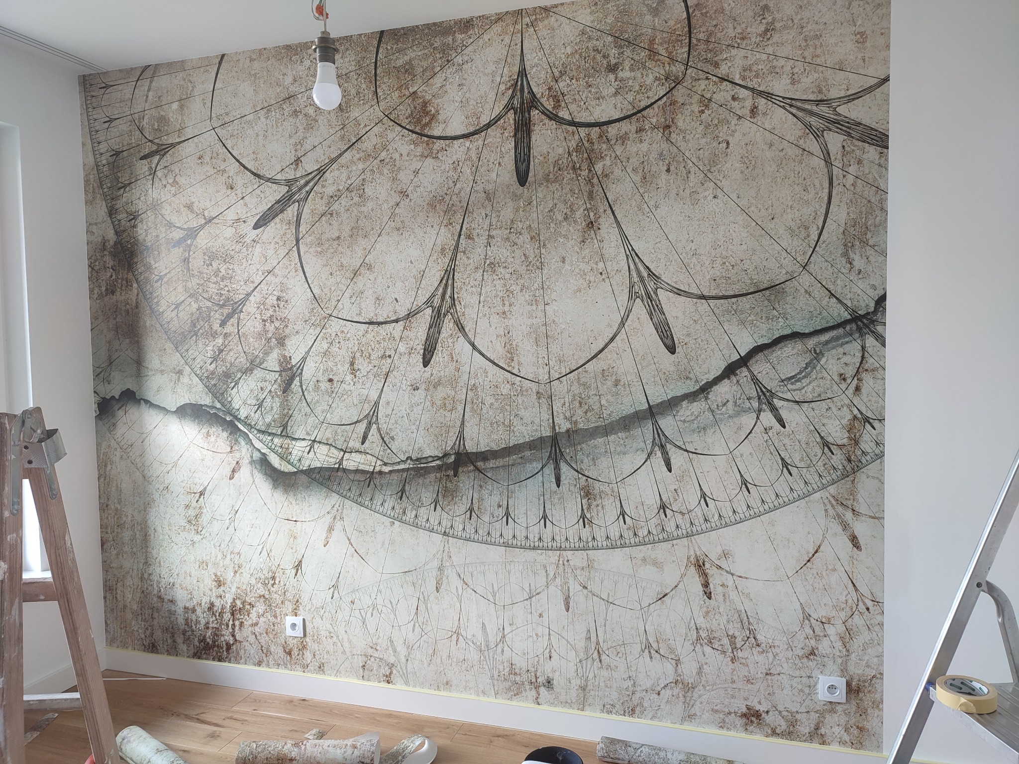 Aplikacja tapety z motywem zegara słonecznego na ścianie, widoczne drabiny i rolki tapety na podłodze, surowa żarówka zwisająca z sufitu.