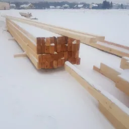 Stosy świeżo przetartej, jasnej tarcicy budowlanej, ułożone na śniegu, z widocznymi oznaczeniami na końcach desek i zabudowaniami w tle.