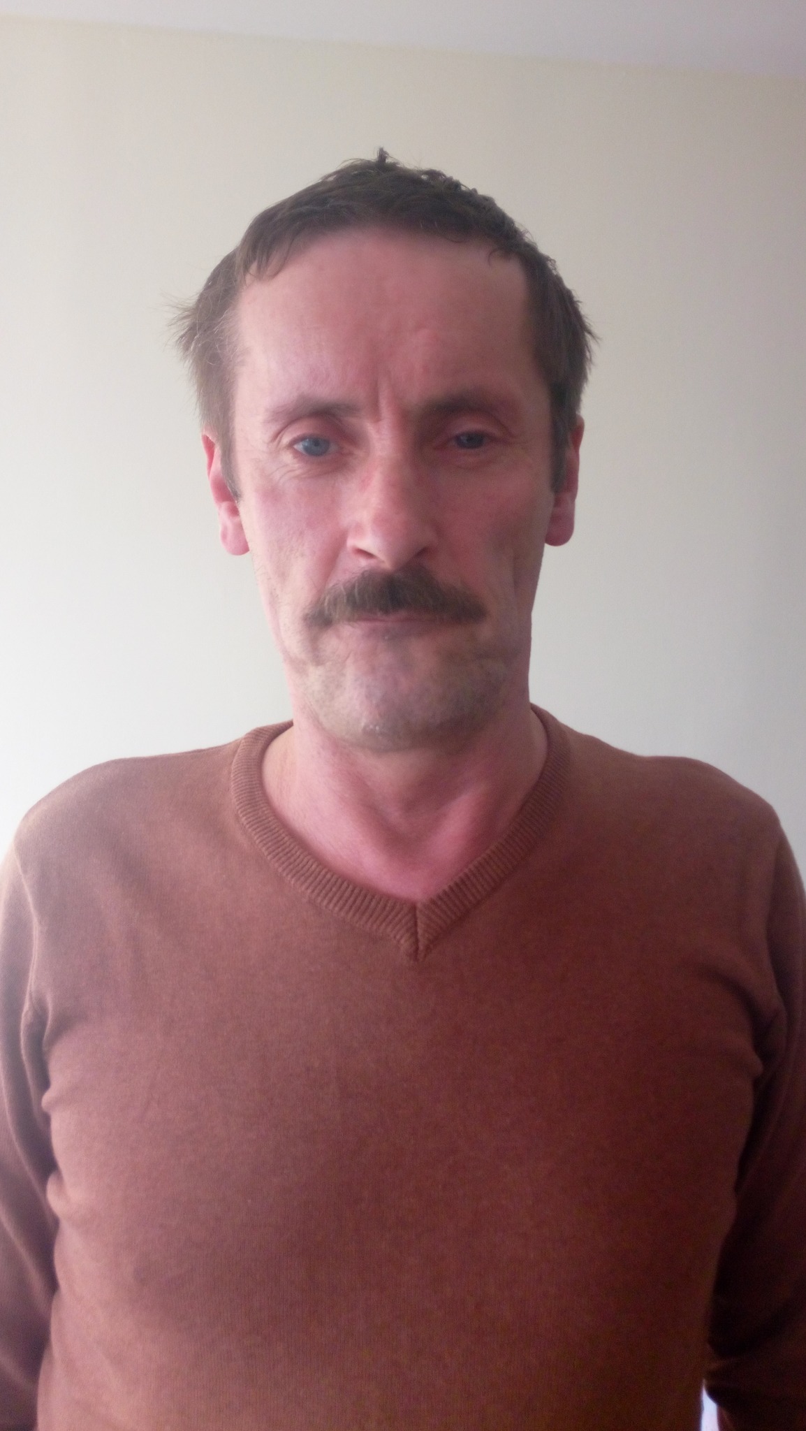 Portret mężczyzny z wąsami w brązowym swetrze na tle jasnej ściany, zdjęcie wykonane w Piasecznie.