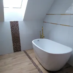Nowoczesna łazienka na poddaszu z wolnostojącą białą wanną, złotą armaturą, wykończona białymi płytkami z dekoracyjnymi paskami mozaiki i oknem dachowym.