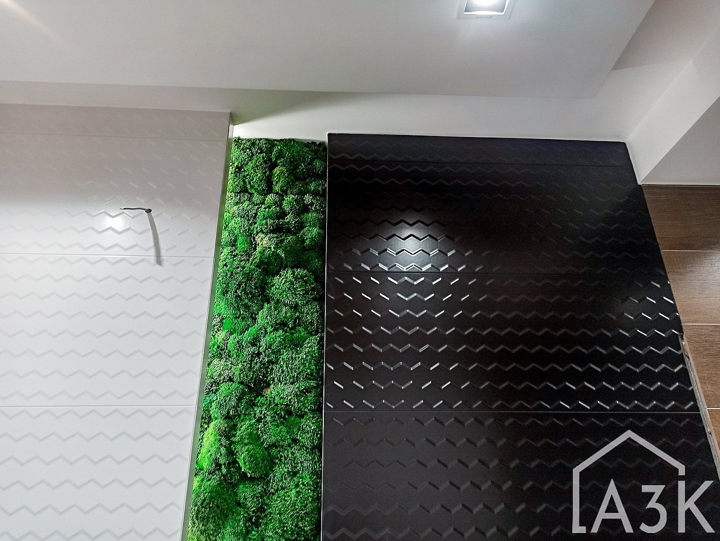 Fragment ściany z dekoracyjnym panelem z mchu, obok białe płytki ze wzorem zygzaków i ciemne płytki z geometrycznym tłoczeniem, oświetlenie sufitowe