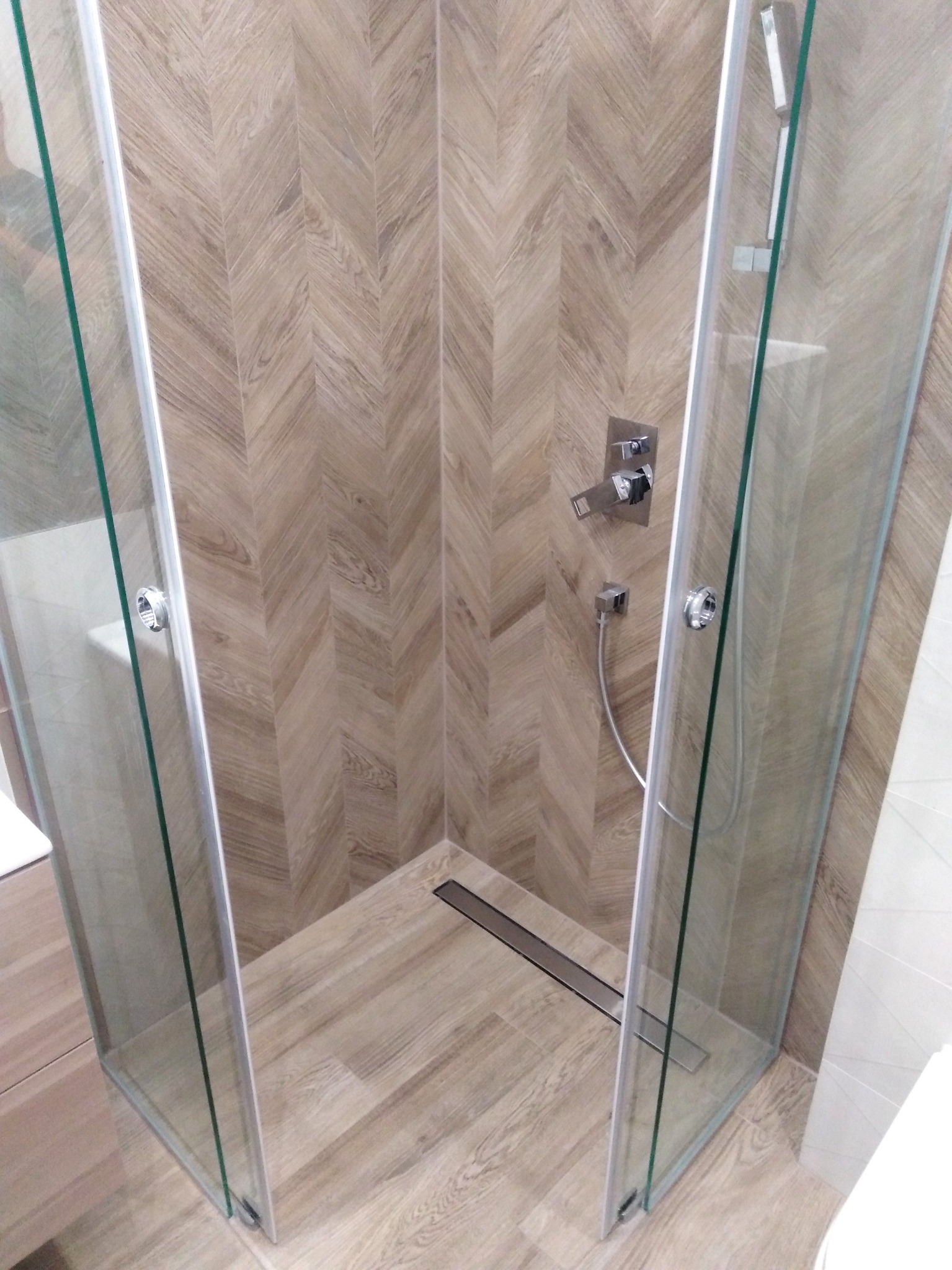 Kabina prysznicowa walk-in z odpływem liniowym, wykończona płytkami imitującymi drewno w jodełkę na ścianach i podłodze, widoczne chromowane elementy armatury.