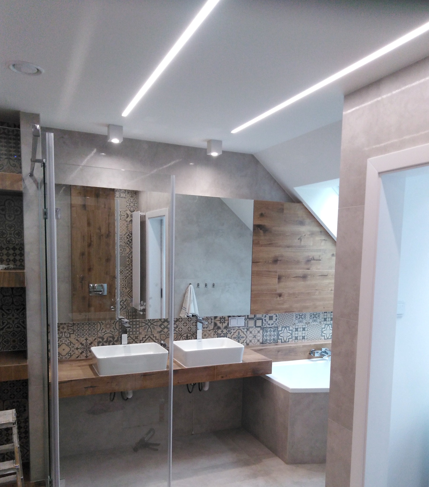 Nowoczesna łazienka z dwoma białymi umywalkami nablatowymi na drewnianym blacie, przeszklona kabina prysznicowa, wanna, płytki patchworkowe na ścianie i oświetlenie LED na suficie.